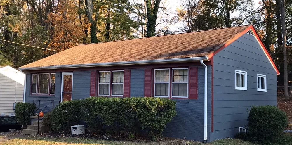 Blue and red exterior home with brown roof and front porch surrounded by shrubs and trees