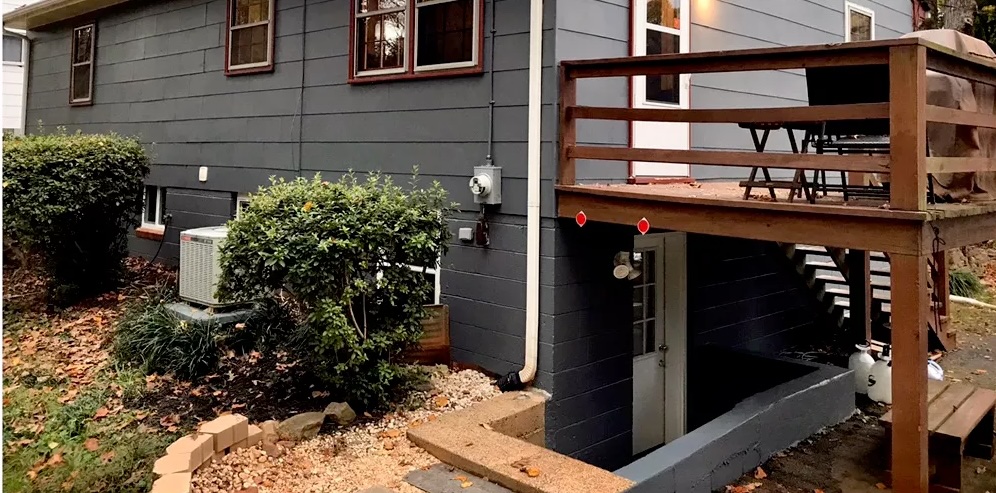 Modern home exterior with dark siding, deck railing, and landscaped front yard with plants