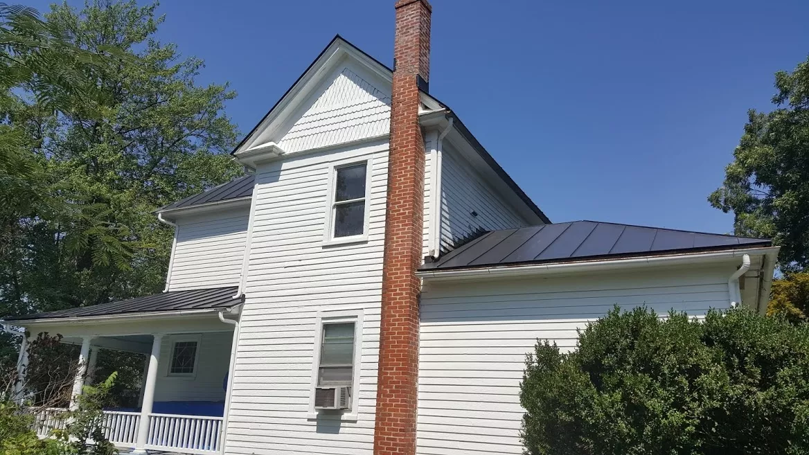 White farmhouse with red brick chimney and metal roof under clear blue sky