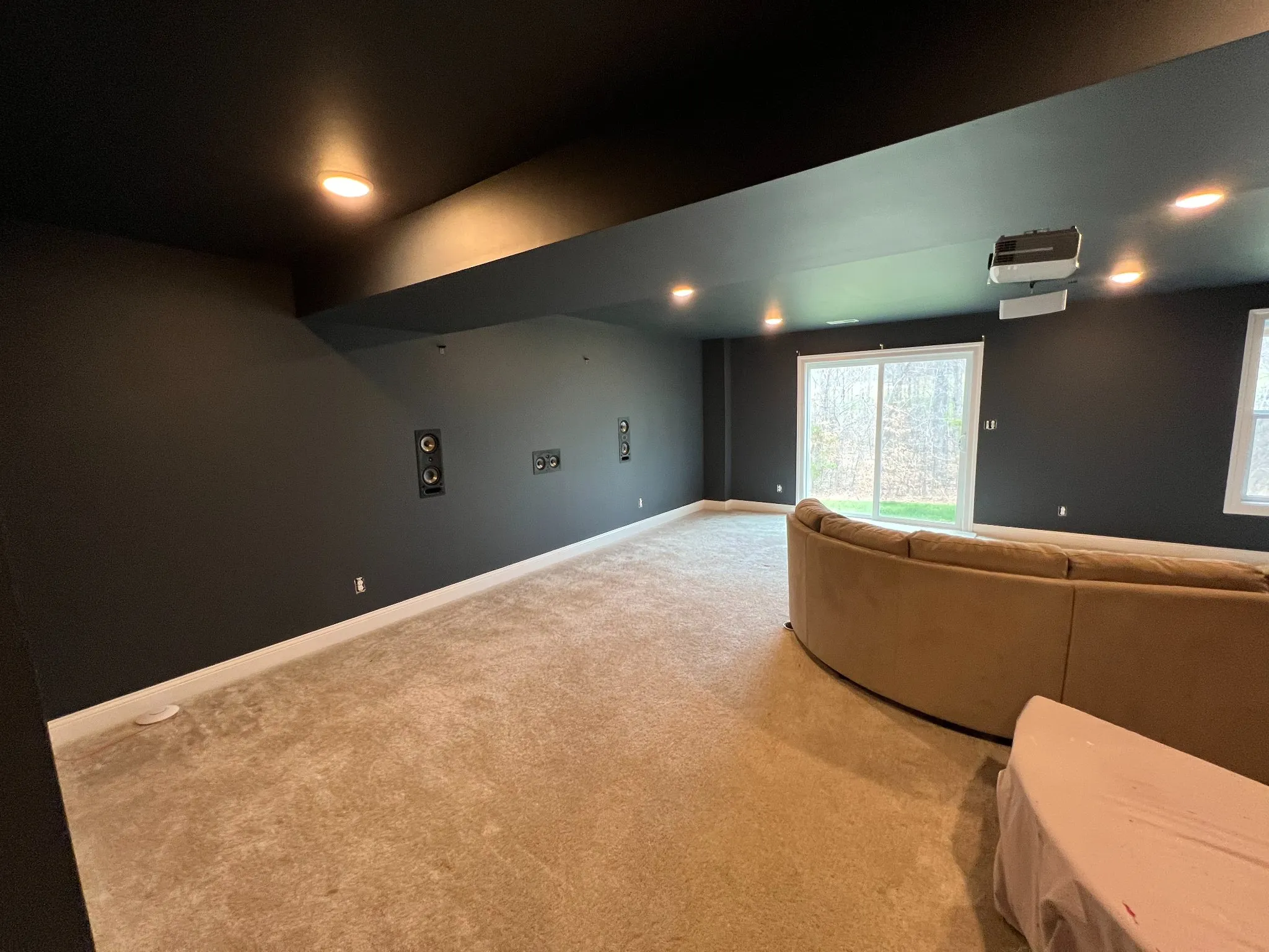 Modern home theater room with dark walls, recessed lighting, leather seating, and glass doors overlooking greenery