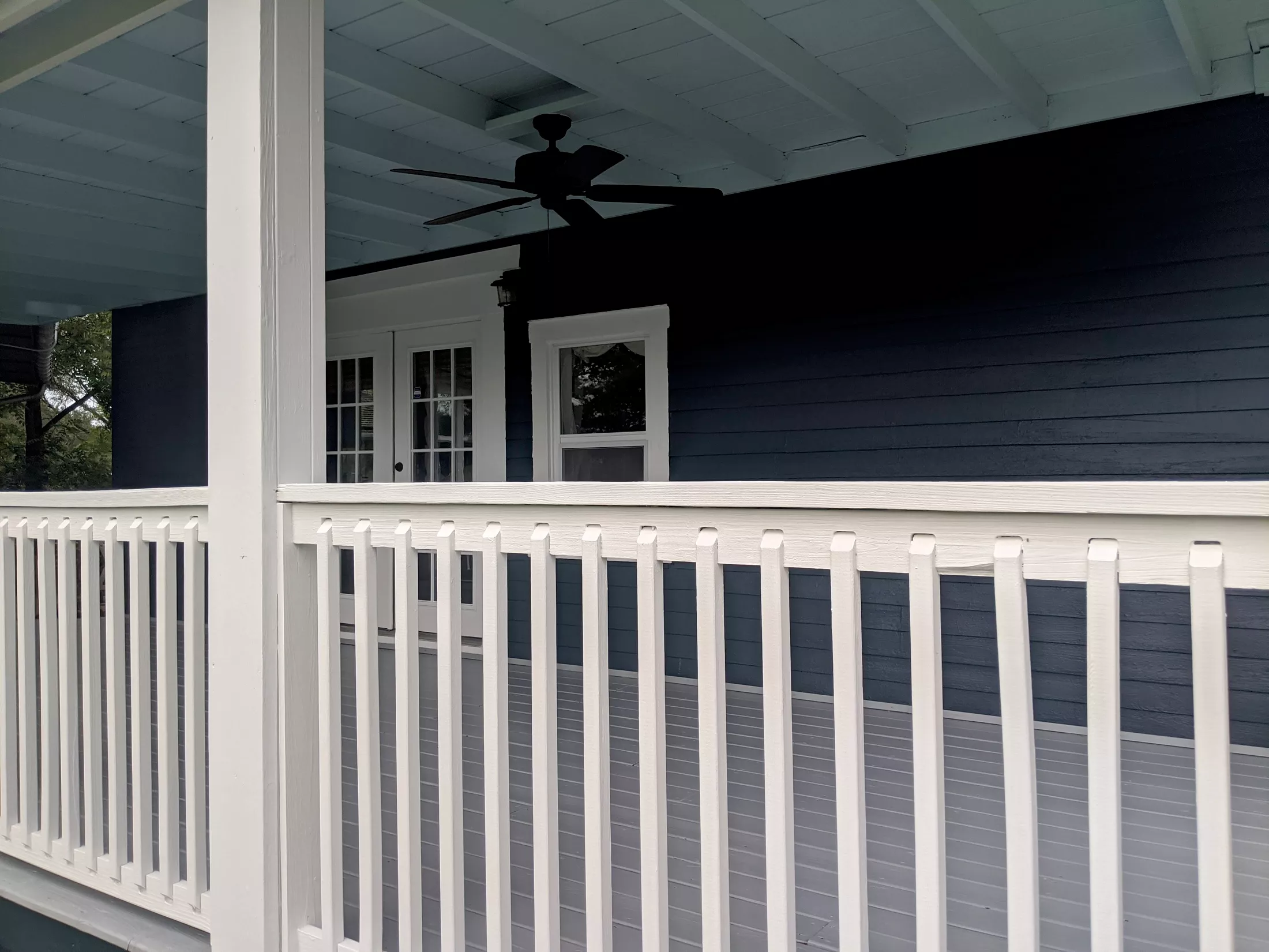 Modern covered porch with white railing, dark siding, ceiling fan, and glass doors