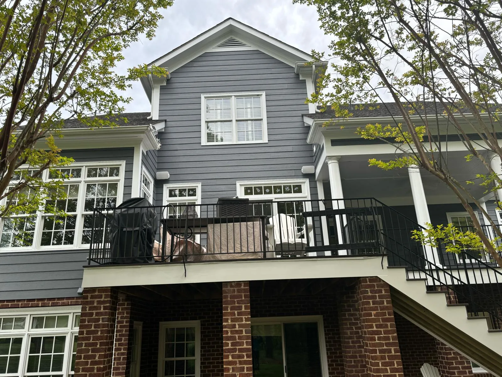 Modern two-story home with gray siding, brick base, black metal deck railing, and white trim surrounded by trees
